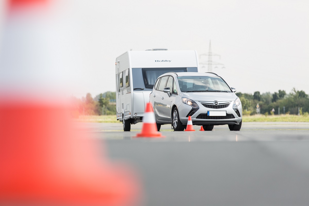 Ein silbernes Auto fährt mit einem Wohnwagen um gestreifte Trainingshütchen.